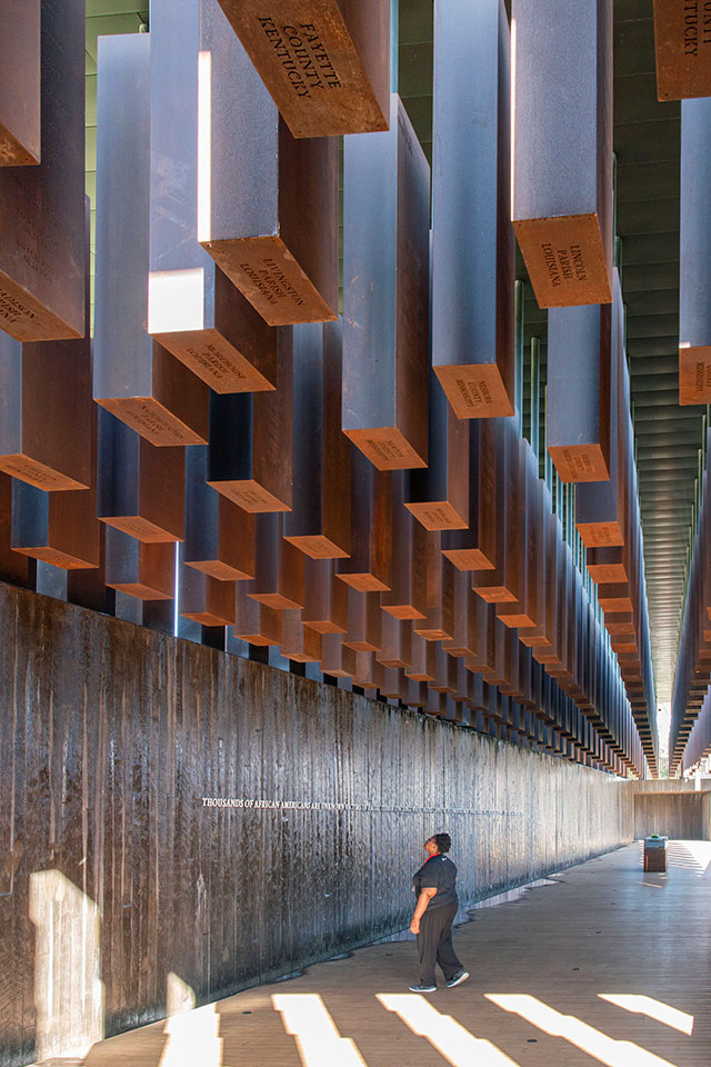 a woman stands under the corten steel monuments at the National Memorial for Peace and Justice.