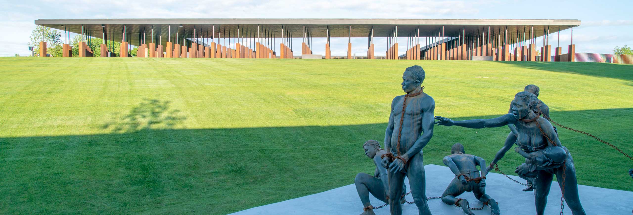 an exterior view of the National Memorial for Peace and Justice. A sculpture of enslaved men and women in chains in the foreground and the memorial building is in the background