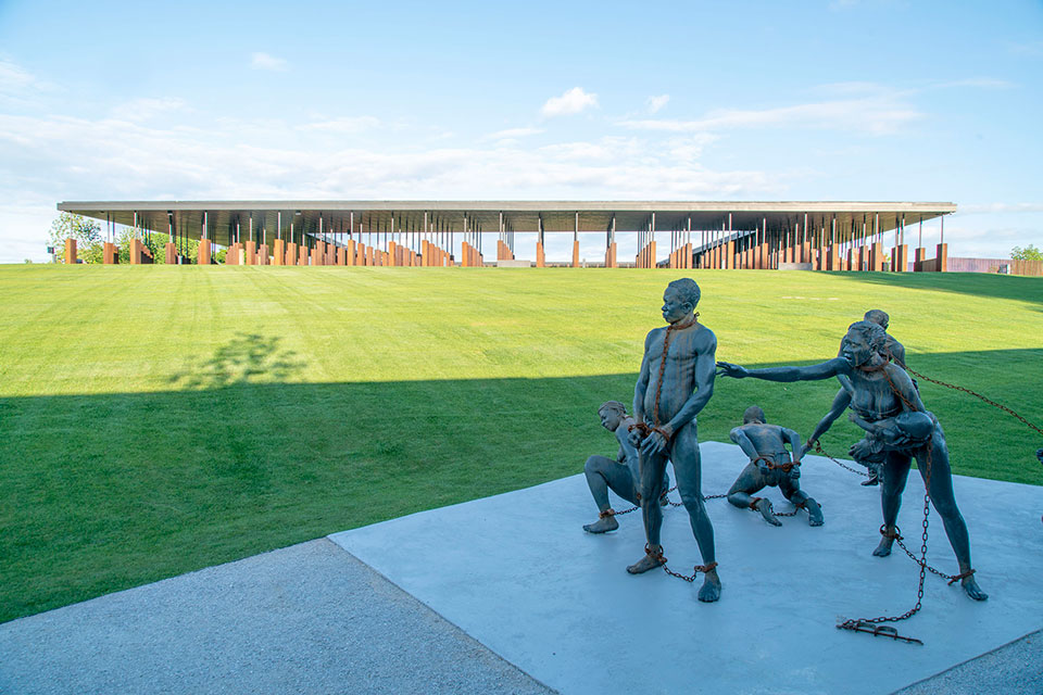 an exterior view of the National Memorial for Peace and Justice. A sculpture of enslaved men and women in chains in the foreground and the memorial building is in the background