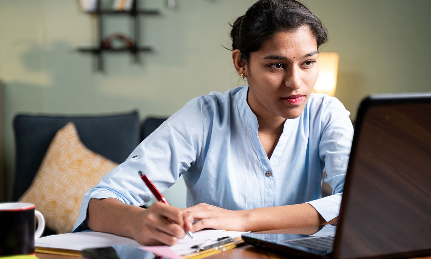 Indian Woman looking at a laptop while taking notes with a pen and paper